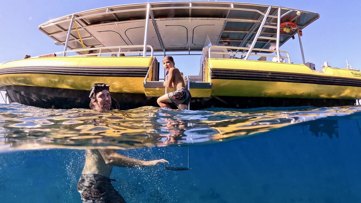 A swimmer beside MAKAI while a young snorkeler climbs the side swim ladder, shot half above and half below the water