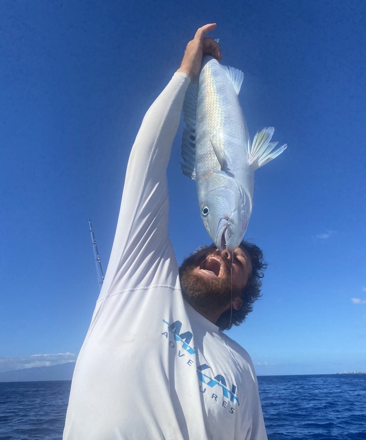 An angler in a Makai Adventures shirt holding a fish overhead against a blue sky